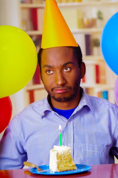 Good looking man wearing blue shirt and yellow party hat sitting by table staring forward, piece of cake placed in front, celebrating alone concept