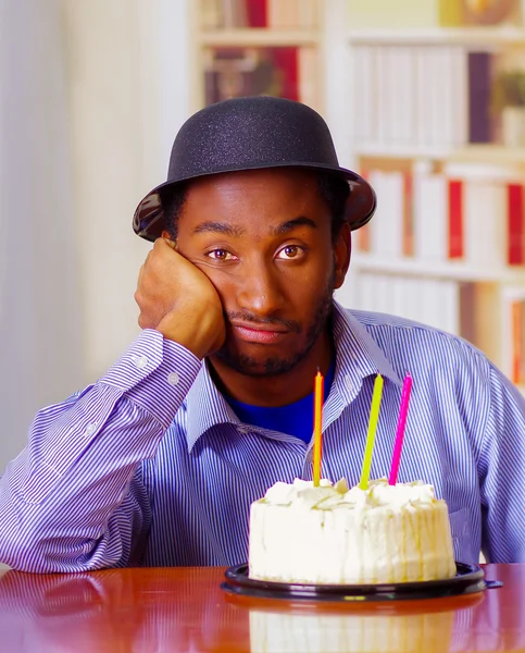 Charming man wearing blue shirt and hat sitting by table with birthday cake in front, looking sad depressed celebrating alone