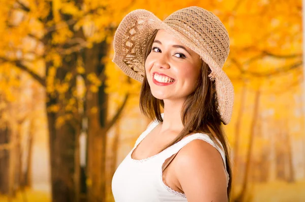 Young attractive woman wearing white top and fashionable hat smiling to camera, forest autumn background