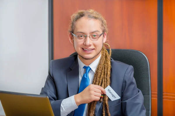 Handsome man with dreads, glasses and business suit sitting by desk placing a paper which has the word ceo written on it in his pocket, young executive concept