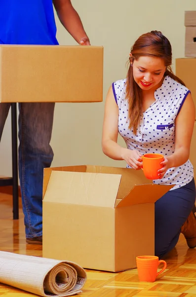 Charming interracial couple working together, woman sitting down unpacking cardboard box while smiling, man standing behind with only legs visible, moving in concept