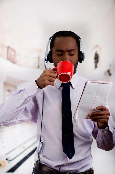 Handsome man wearing headphones with microphone, white striped shirt and tie, drinking from coffee mug, business concept