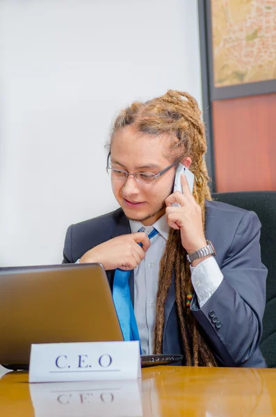 Handsome man with dreads and business suit sitting by desk talking on mobile phone, young manager concept