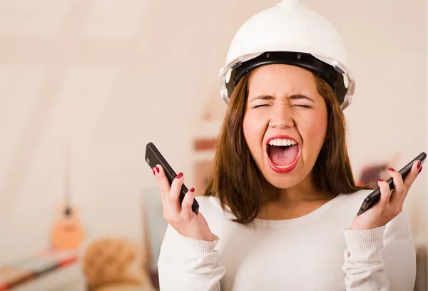 Young woman wearing construction helmet facing camera, holding two mobile phones and screaming out in frustration