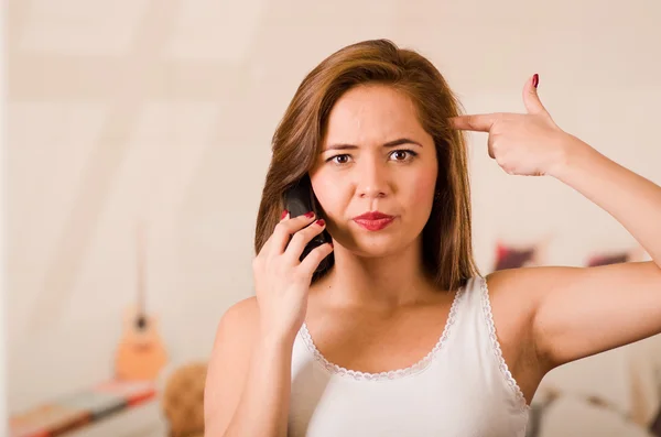 Young woman wearing white top facing camera while interacting frustration talking on phone, making gun with fingers pointing to her own head