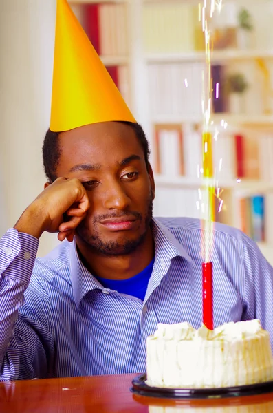 Sad man wearing blue shirt and hat sitting by table with cake in front, single candle burning, looking bored depressed, celebrating alone concept