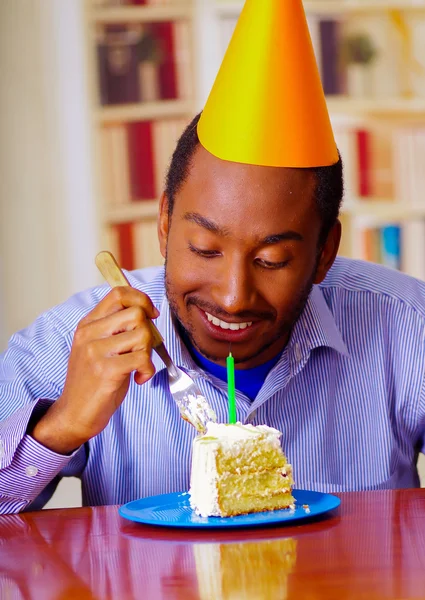 Charming man wearing blue shirt and hat sitting by table with piece of cake in front, looking happy, celebrating alone concept