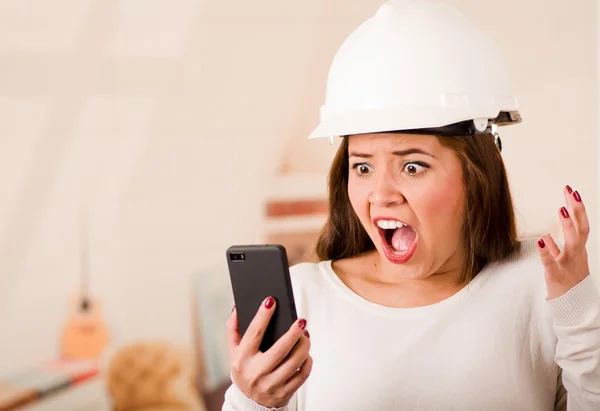 Young woman wearing construction helmet looking at mobile phone with stressed facial expression