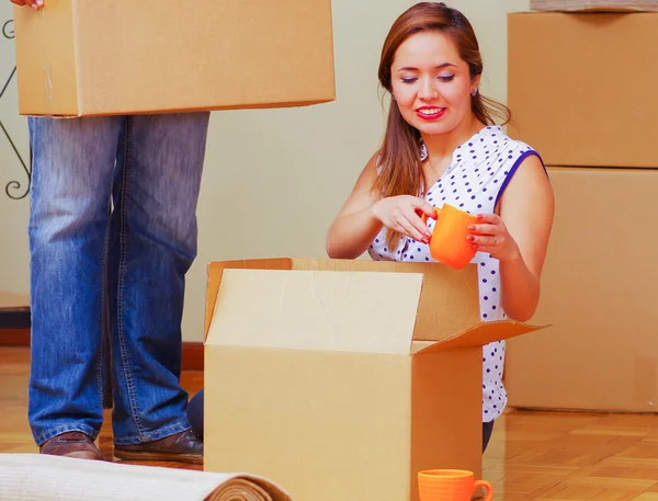 Charming interracial couple working together, woman sitting down unpacking cardboard box while smiling, man standing behind with only legs visible, moving in concept
