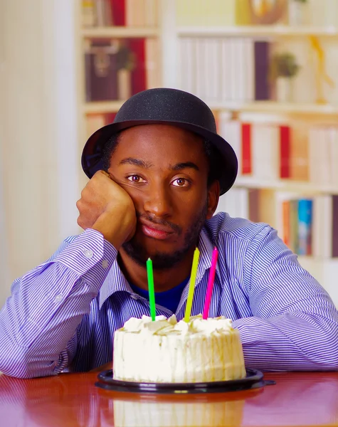 Charming man wearing blue shirt and hat sitting by table with birthday cake in front, looking sad depressed celebrating alone