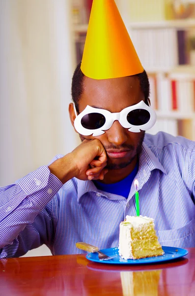 Good looking man wearing blue shirt, white spectacular sunglasses and yellow party hat sitting by table staring at piece of cake in front, celebrating alone concept