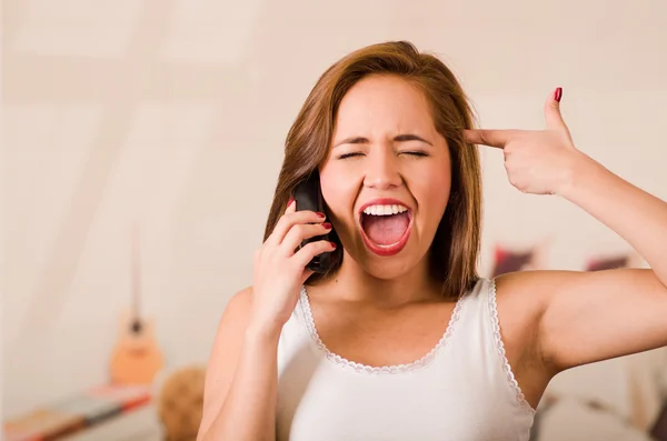 Young woman wearing white top facing camera while interacting frustration talking on phone, making gun with fingers pointing to her own head