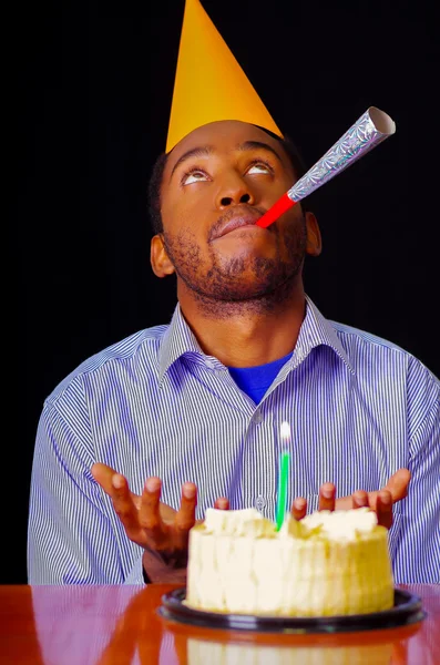 Good looking man wearing blue shirt and hat sitting by table with cake in front, single candle burning, blowing party horn facing camera, celebrating alone concept
