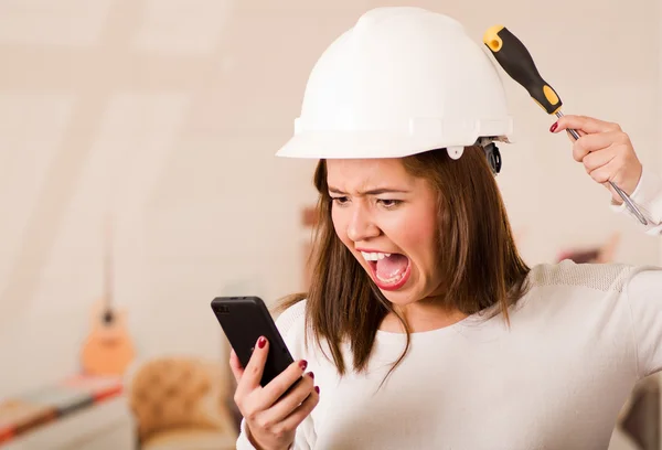 Young woman wearing construction helmet facing camera, looking at mobile screen, simulating smashing tool against her own head in frustration