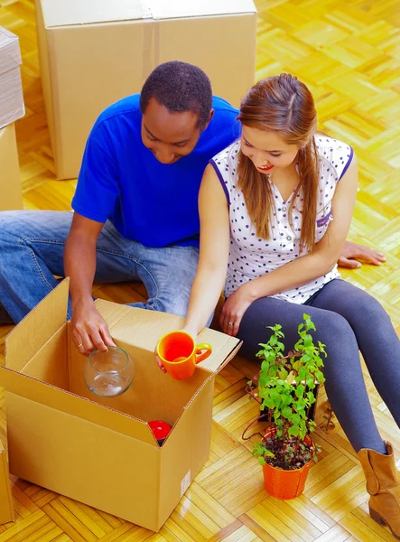 Charming interracial couple working together, sitting down and unpacking cardboard box while smiling, moving in concept