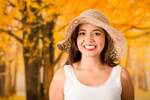 Young attractive woman wearing white top and fashionable hat smiling to camera, forest autumn background
