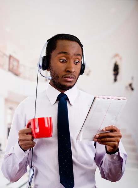 Handsome man wearing headphones with microphone, white striped shirt and tie, holding coffee mug, surprised facial expression, business concept