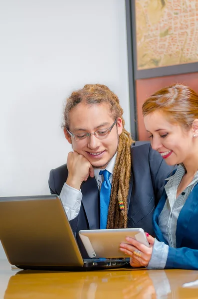 Handsome young man wearing formal suit sitting down with pretty female co-worker looking at screen and discussing between each other, office manager concept