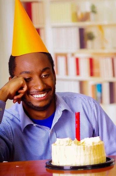 Good looking man wearing blue shirt and hat sitting by table with cake in front, single candle burning, skeptically smiling, celebrating alone concept