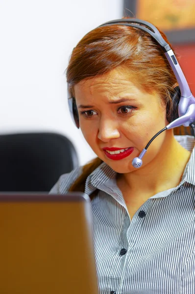 Young attractive woman wearing office clothes and headset sitting by desk looking at computer screen, working with positive facial expression