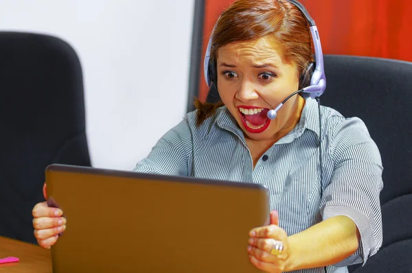Young attractive woman wearing office clothes and headset sitting by desk looking at computer screen, grabbing laptop, upset body language screaming out loud