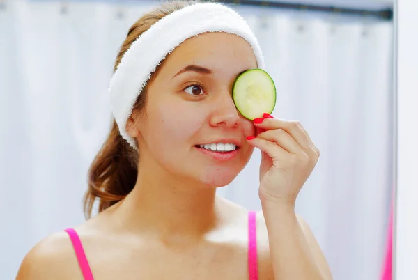 Attractive young woman wearing pink top and white headband, covering one eye with slice of cucumber used for skin treatment, looking in mirror smiling
