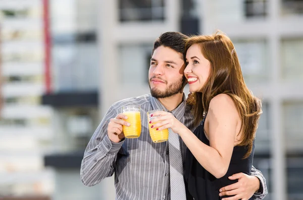Young attractive couple wearing formal clothes standing on rooftop holding glass with yellow drink, posing, smiling and embracing, city buildings background