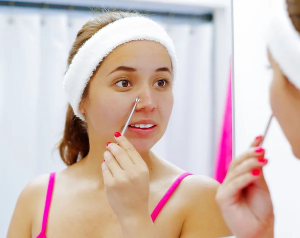 Attractive young woman wearing pink top and white headband, squeezing nose with flat tweezers, looking in mirror smiling