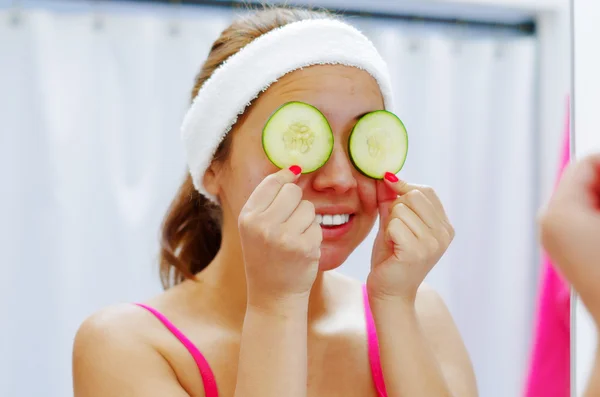 Attractive young woman wearing pink top and white headband, covering both eyes with slices of cucumber used for skin treatment, looking in mirror smiling