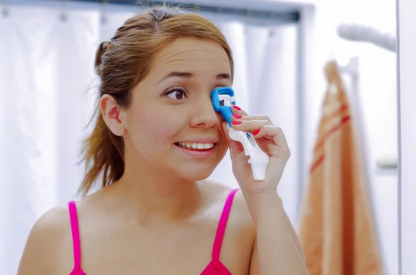 Attractive young woman wearing pink top and white headband, using eyelash curler tool on left eye, looking in mirror smiling