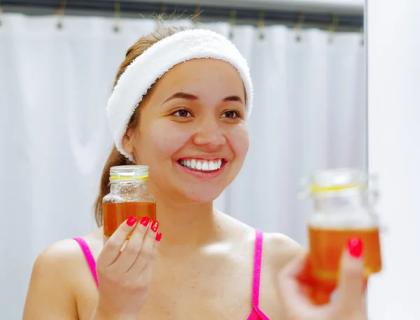 Attractive young woman wearing pink top and white headband, holding up jar of honey, looking in mirror smiling