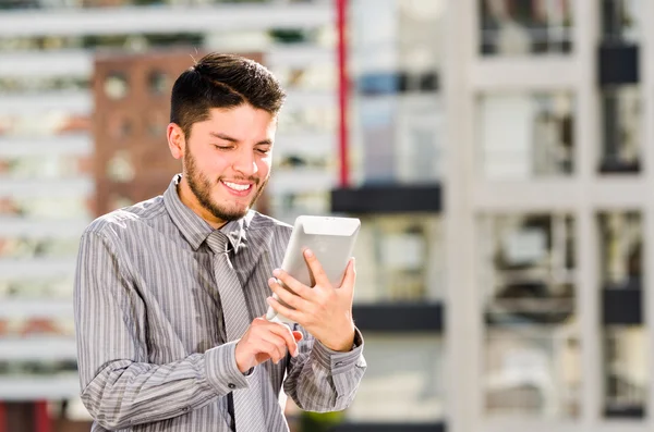 Young handsome man wearing shirt and tie standing on rooftop, holding tablet staring at screen, city buildings background