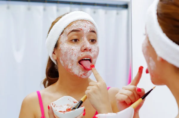 Attractive young woman wearing pink top and white headband with cream on face, holding up finger nearly touching tongue, simulating eating, looking in mirror