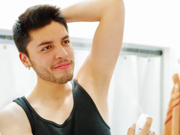 Handsome young man wearing black singlet top looking in mirror, applying deodorant during morning routine concept, sligthly smiling
