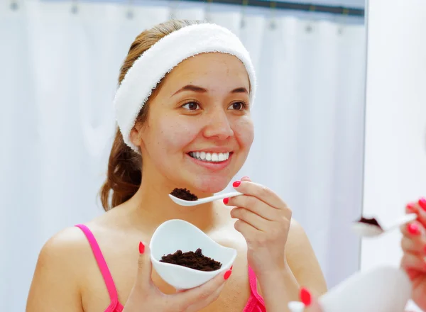 Attractive young woman wearing pink top and white headband, holding up bowl of dark chocolate, looking in mirror smiling