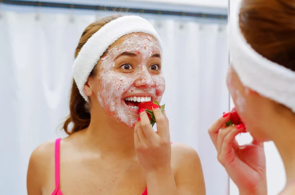 Attractive young woman wearing pink top and white headband with cream on face, eating strawberry, looking in mirror smiling
