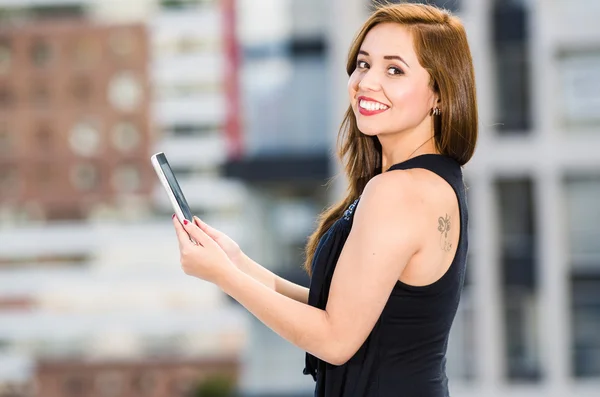 Young attractive woman wearing black dress standing on rooftop, holding tablet, smiling to camera, city buildings background