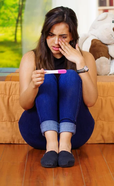 Pretty young brunette woman sitting on floor holding pregnancy home test, crying wiping tears, looking emotional, garden window background