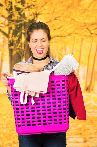 Attractive brunette wearing casual clothes while carrying pink plastic basket with different products, smiling posing for camera, autumn forest background