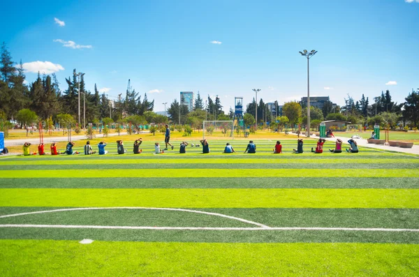 QUITO, ECUADOR - 8 AUGUST, 2016: Row of people stretching seated on football field located in inner city park La Carolina, artificial green grass surface, buildings visible background, beautiful sunny