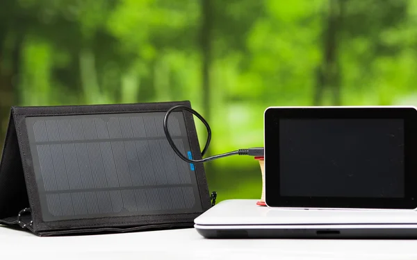 Portable solar charger sitting on white desk surface connected to tablet, modern technology concept, window garden background