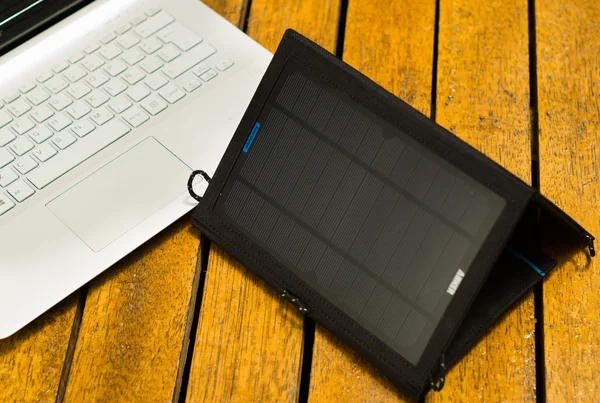 Portable solar charger sitting on wooden surface next to laptop computer, as seen from above, modern technology concept