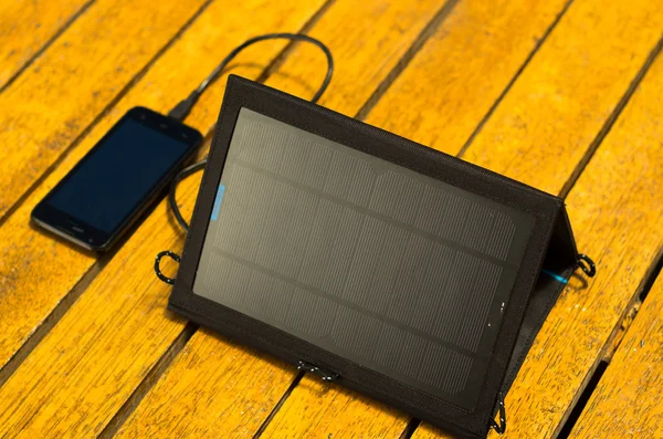 Portable solar charger sitting on wooden surface next to mobile phone, as seen from above, modern green technology concept