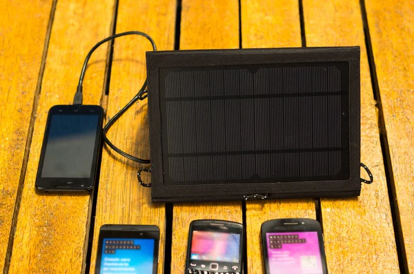 Portable solar charger sitting on wooden surface next to four mobile phones, as seen from above, modern green technology concept