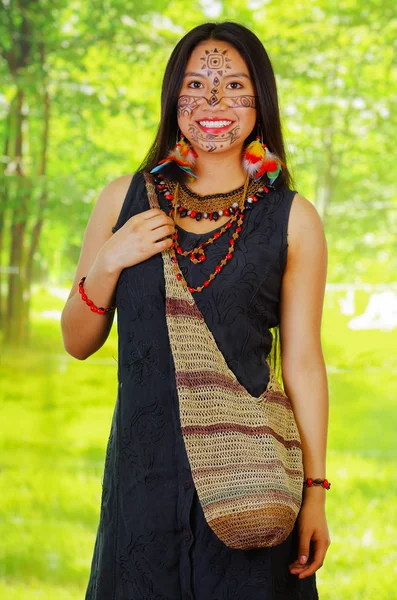 Amazonian exotic woman with facial paint and black dress, natural bag hanging across upper body, posing for camera, forest background