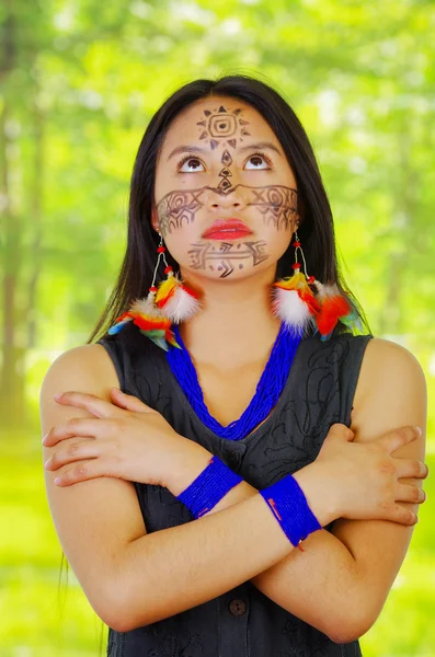 Portrait amazonian exotic woman with facial paint and black dress, posing proudly for camera, arms crossed across upper body, forest background.