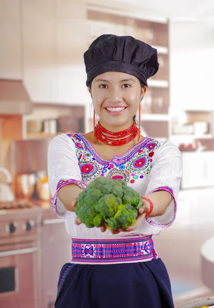Young woman chef wearing traditional andean blouse, black cooking hat, holding broccoli up showing to camera and smiling, kitchen background