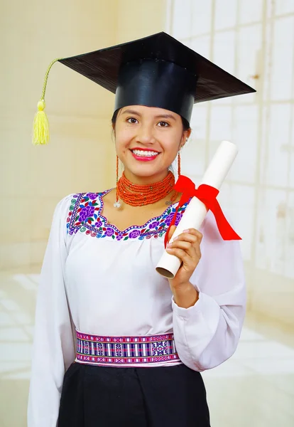 Young female student wearing traditional blouse and graduation hat, holding rolled up diploma, smiling proudly for camera