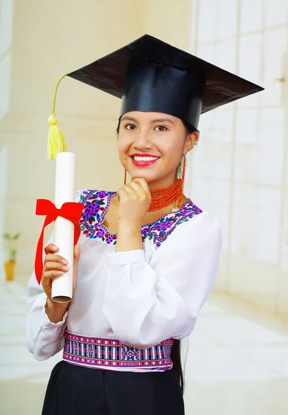 Young female student wearing traditional blouse and graduation hat, holding rolled up diploma, smiling proudly for camera