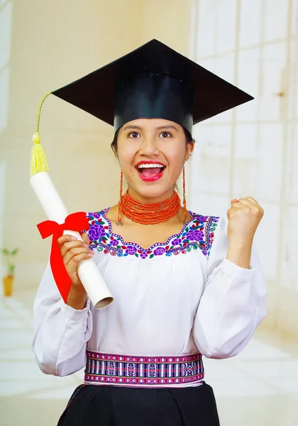Young female student wearing traditional blouse and graduation hat, holding rolled up diploma, smiling proudly for camera